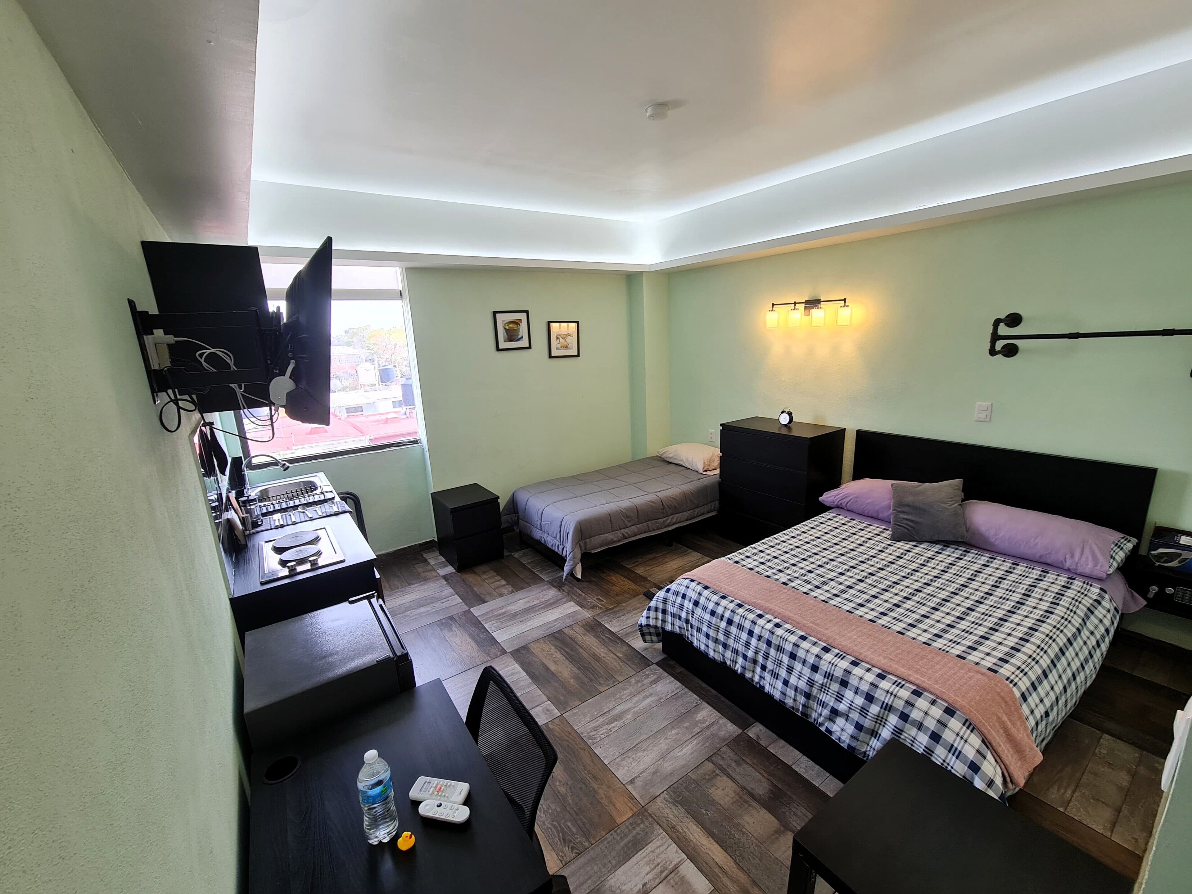 Un dormitorio con cama, escritorio, silla, mesita, televisor y ventana.