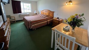 Habitación de hotel con cama, escritorio, silla, televisor, lámpara y arreglo floral.