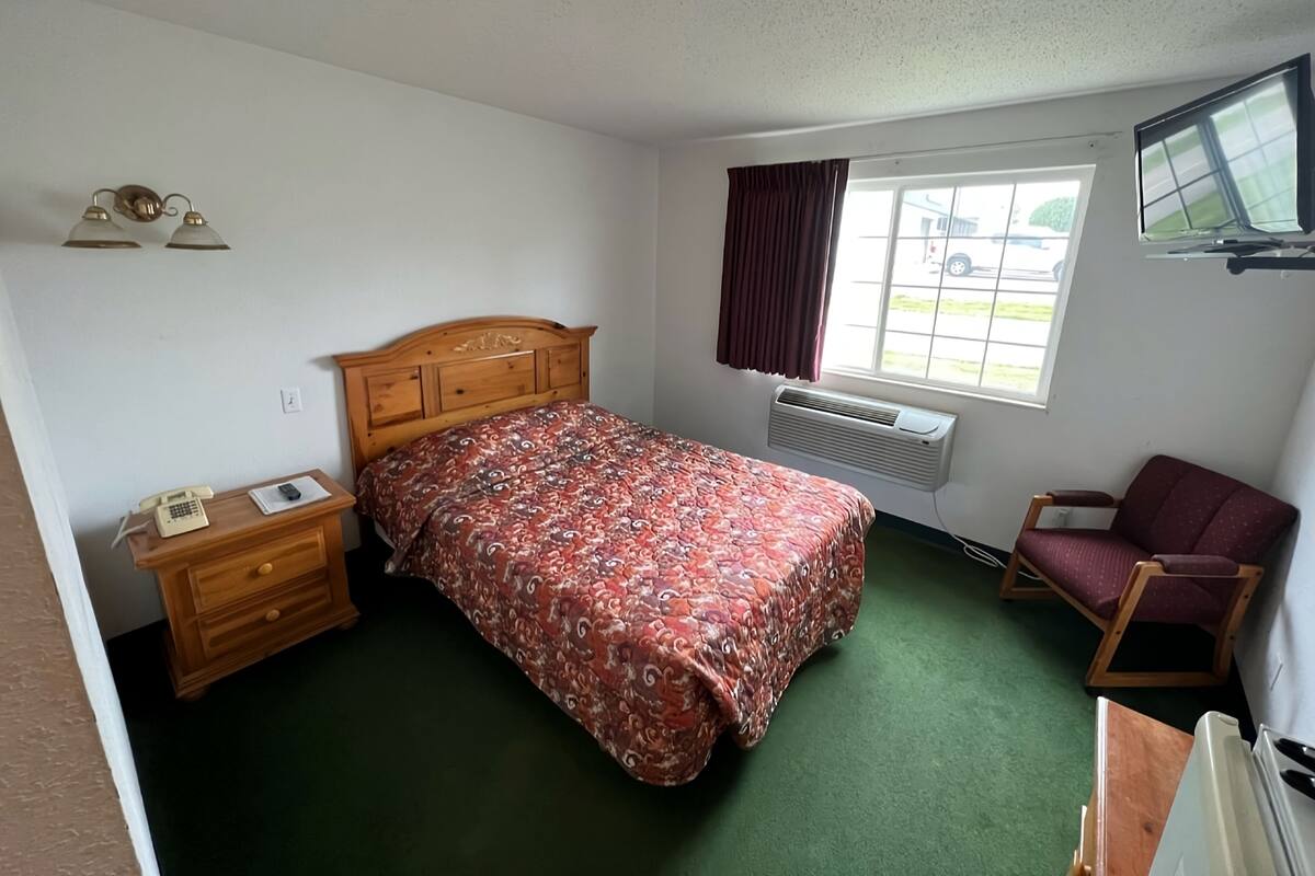 A bedroom with a wooden bed, a nightstand with a telephone, a chair, a window with curtains, and a wall-mounted TV.