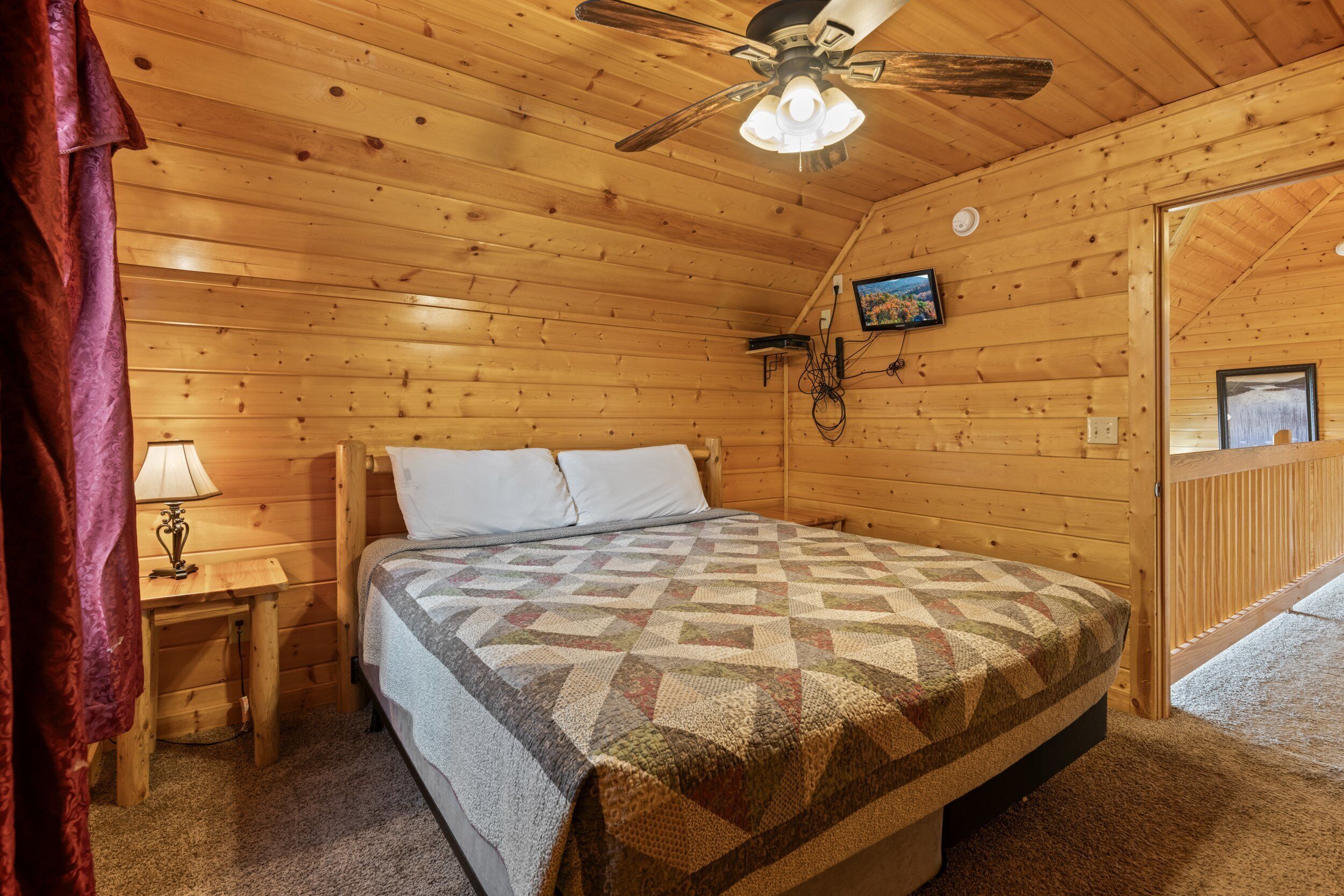 A wooden cabin bedroom with a bed, a nightstand, a lamp, a ceiling fan, and a door leading to another room.