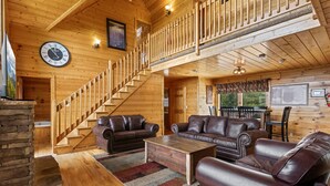Ein großzügiges Holzhütten-Interieur mit einer Treppe, zwei Ledersofas, einem Couchtisch und einer Wanduhr.