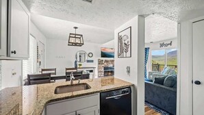 A compact kitchen with granite countertops, white cabinetry, and a dishwasher. There is a dining area with a table and chairs, and a living room with a sofa and a view of the outdoors. - Mountain View Condo w/ Indoor Pool & Hot Tub (Gatlinburg)