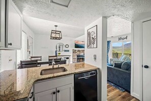 Eine kompakte Küche mit Granit-Arbeitsplatten, weißen Schränken und einem Geschirrspüler. Es gibt einen Essbereich mit Tisch und Stühlen sowie ein Wohnzimmer mit einer Couch und Blick ins Freie.