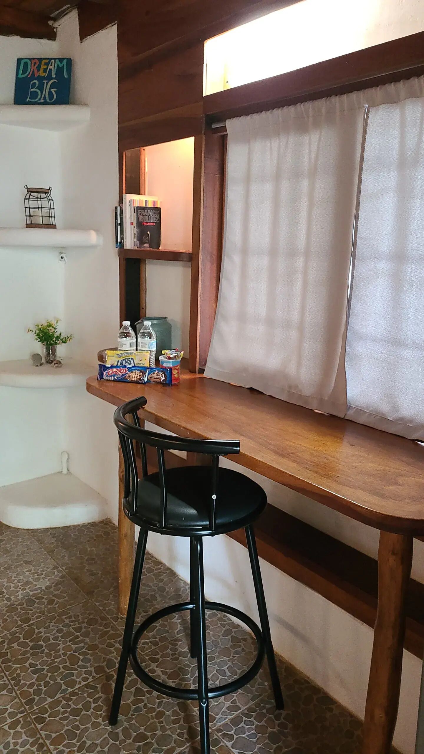 Una mesa de comedor de madera con una silla negra, una estantería con libros y un letrero que dice 'Dream Big', y una ventana con cortinas.