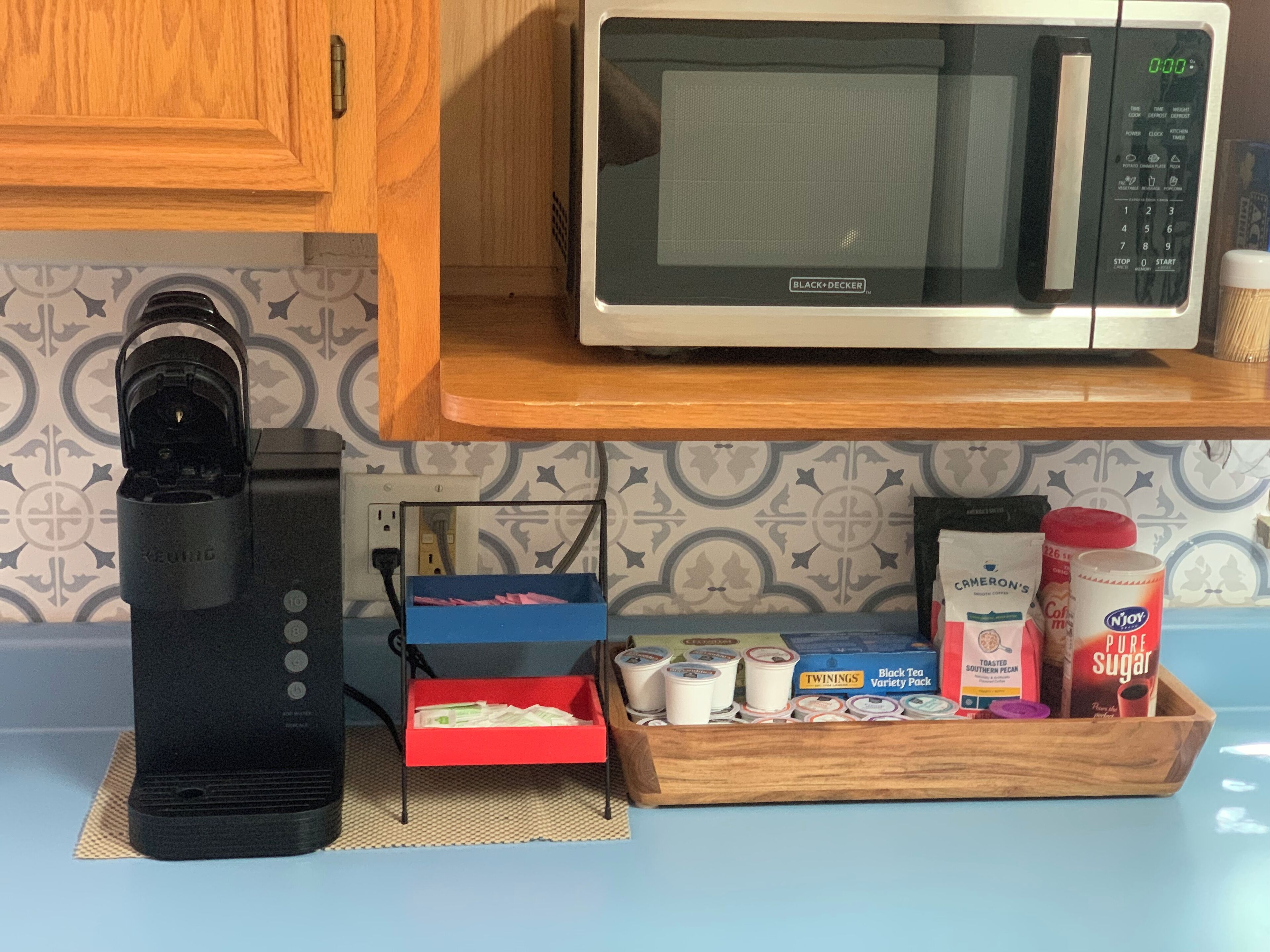 A kitchen with a microwave, a Keurig coffee maker, a coffee maker, and a tray with various coffee pods and sugar.