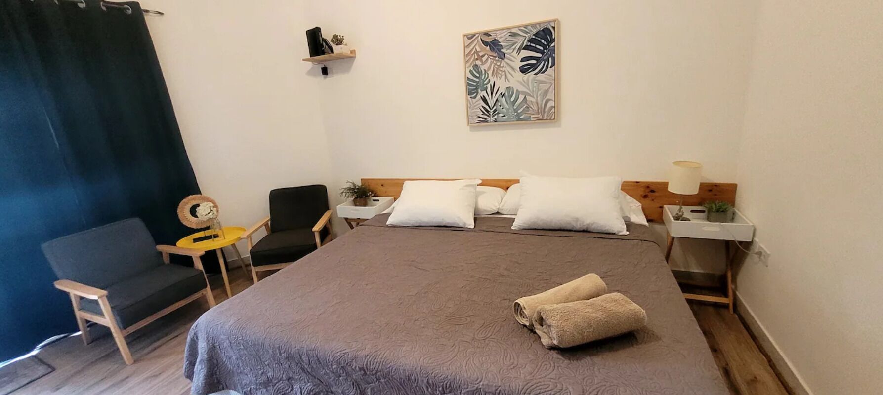 Habitación de hotel con una cama grande, dos mesitas de noche, una silla, una pequeña mesa con lámpara y un estante en la pared con objetos decorativos.