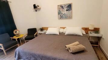 Habitación de hotel con una cama grande, dos mesitas de noche, una silla, una pequeña mesa con lámpara y un estante en la pared con objetos decorativos.