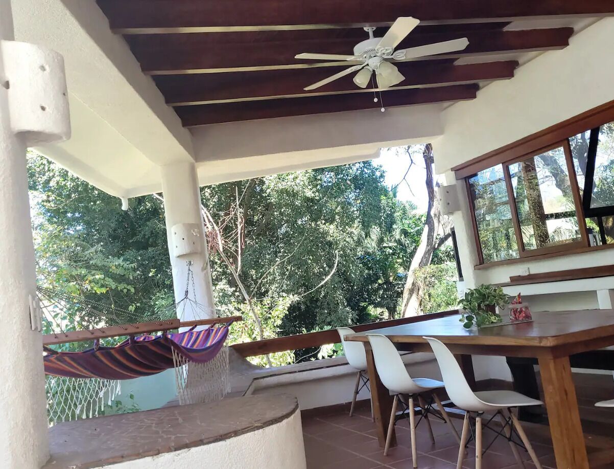 A covered outdoor area with a wooden table and chairs, a hammock, and a ceiling fan.