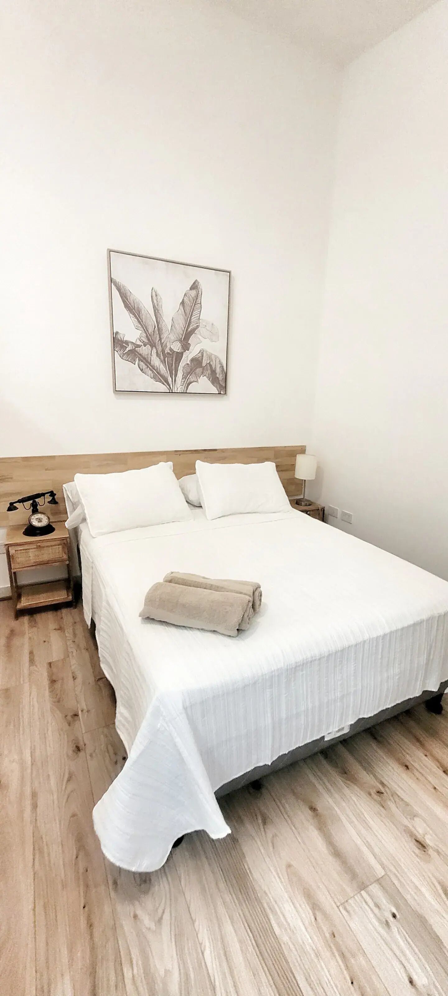 A neatly made bed with white linens and a beige throw blanket, a wooden headboard, a bedside table with a lamp, and a framed botanical print on the wall.