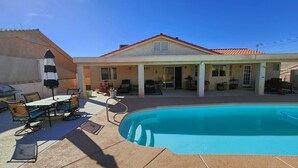 Un área junto a la piscina con un porche cubierto, mobiliario exterior y un edificio con tejado de baldosas.