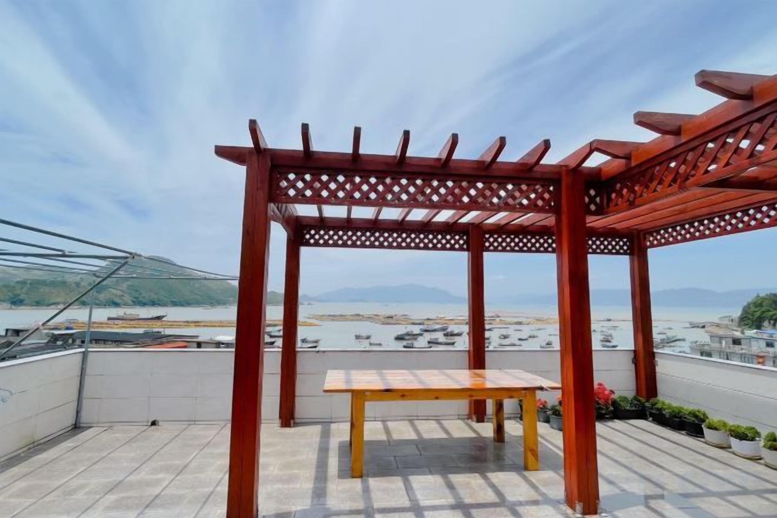 A red wooden pergola with a table on a rooftop terrace with a view of the sea and boats.