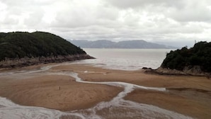 A beach with a sandy shore, a small water channel, and a forested hill in the background.