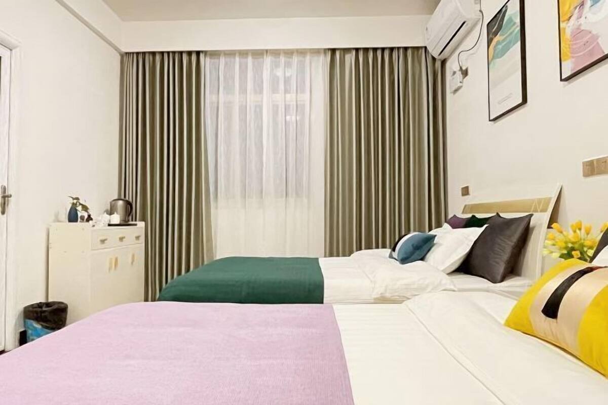 A hotel room with a bed, a desk, a chair, a window with curtains, and an air conditioning unit.