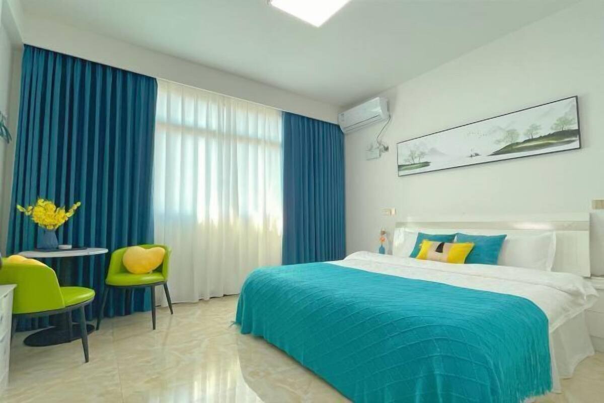 A hotel room with a bed, a desk with chairs, a window with curtains, and a painting on the wall.