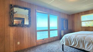 Une chambre avec des murs en bois, une grande fenêtre donnant sur la mer, un lit et une commode.