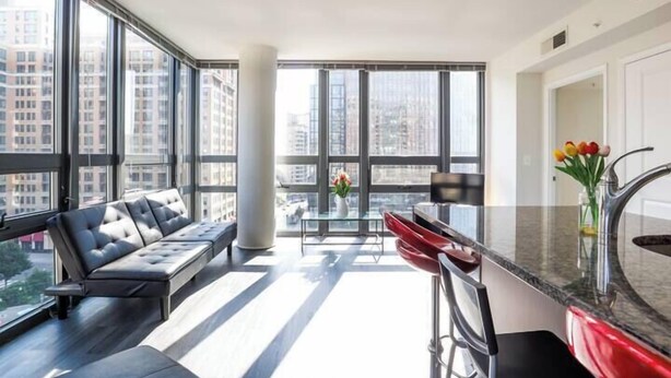 A modern apartment with a large window, a sofa, a kitchen area, and a view of the city. - Pentagon Luxe Apartments (Arlington)