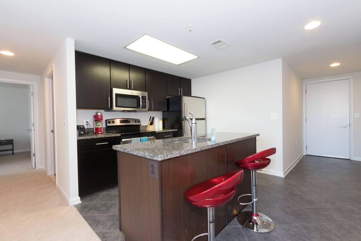 A modern kitchen with dark cabinets, stainless steel appliances, and a granite countertop. There are two red bar stools and a small island with a sink.