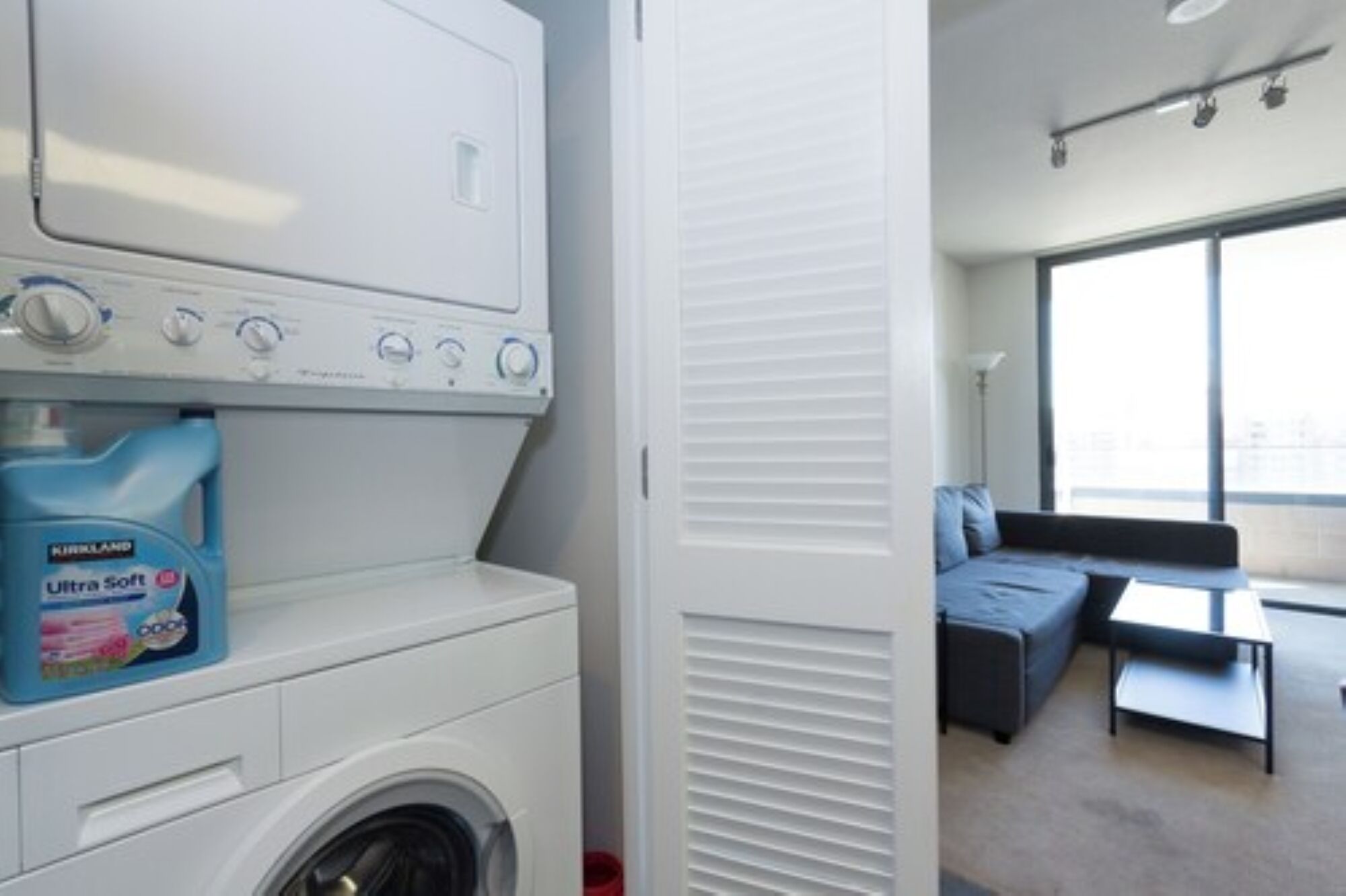 Un espace buanderie comprenant un lave-linge et un sèche-linge à chargement frontal, une bouteille de lessive bleue et une porte de placard coulissante.