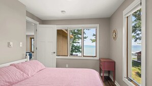 A bedroom with a large bed, a window with a view of the sea, a small desk, and a door leading to another room. - Torch Ever Blue | SandBar | Waterfront (Rapid City)