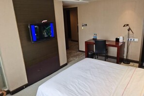 A hotel room with a bed, a desk, a chair, a TV mounted on the wall, and a lamp.