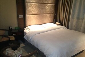A hotel room with a large bed, a wooden headboard, a bedside table with a lamp, and a chair with a cushion.