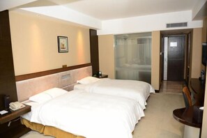 A hotel room with two beds, a desk, a chair, a TV, and a bathroom with a glass door.