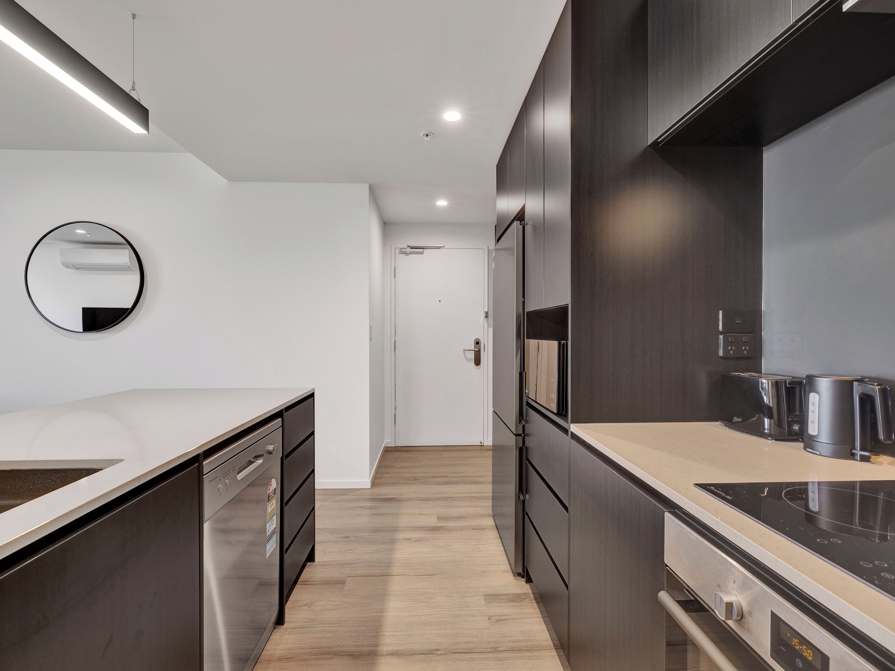 Una cocina moderna con horno integrado, microondas y tostadora. Tiene suelo de madera y encimera blanca.