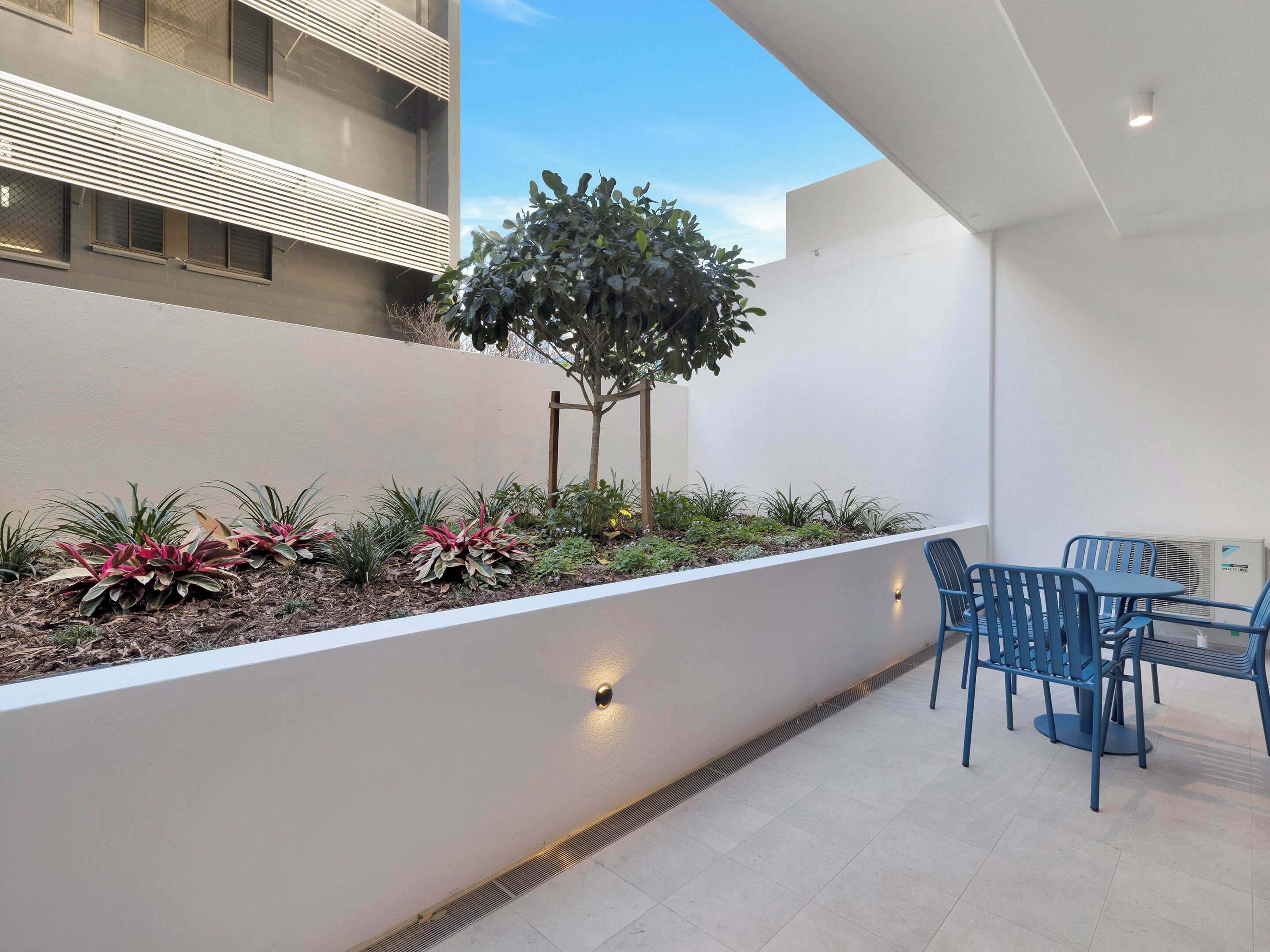 A modern outdoor patio with a seating area, a planter with various plants, and a building with a white facade and a tree.