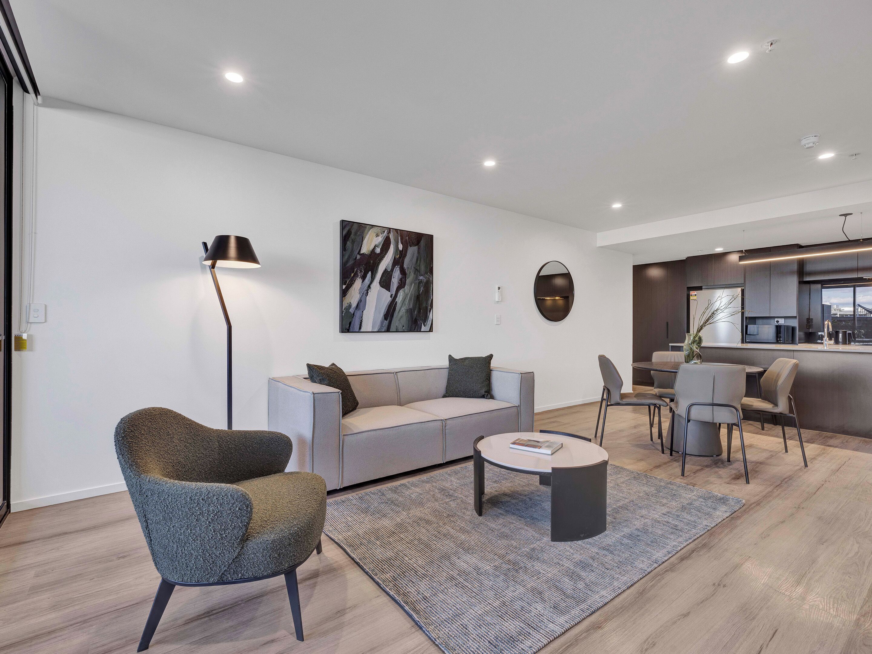 Una sala de estar moderna con un sofá gris, una mesa de centro redonda y un sillón gris. Hay una zona de cocina con una mesa de comedor y sillas.