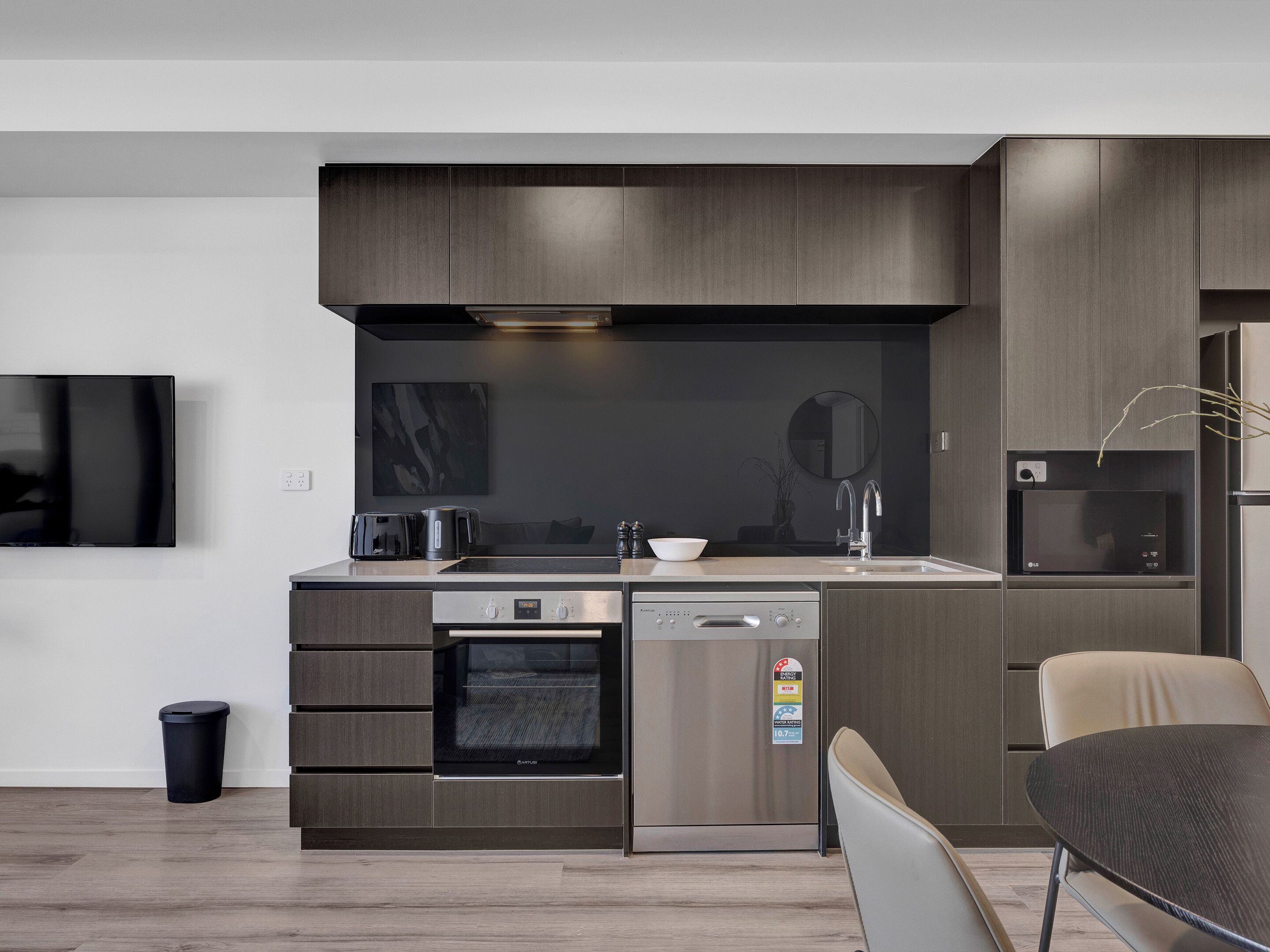 A modern kitchen with dark wood cabinets, a built-in oven, a dishwasher, a microwave, and a large window above the sink.