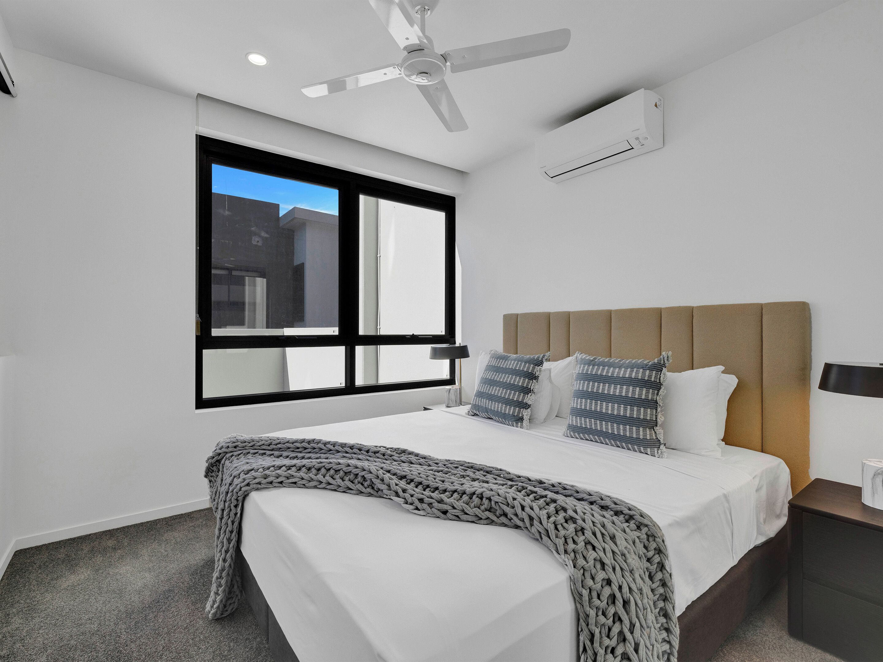 A modern hotel room with a bed, a ceiling fan, an air conditioning unit, a window with a view, and a bedside table with a lamp.