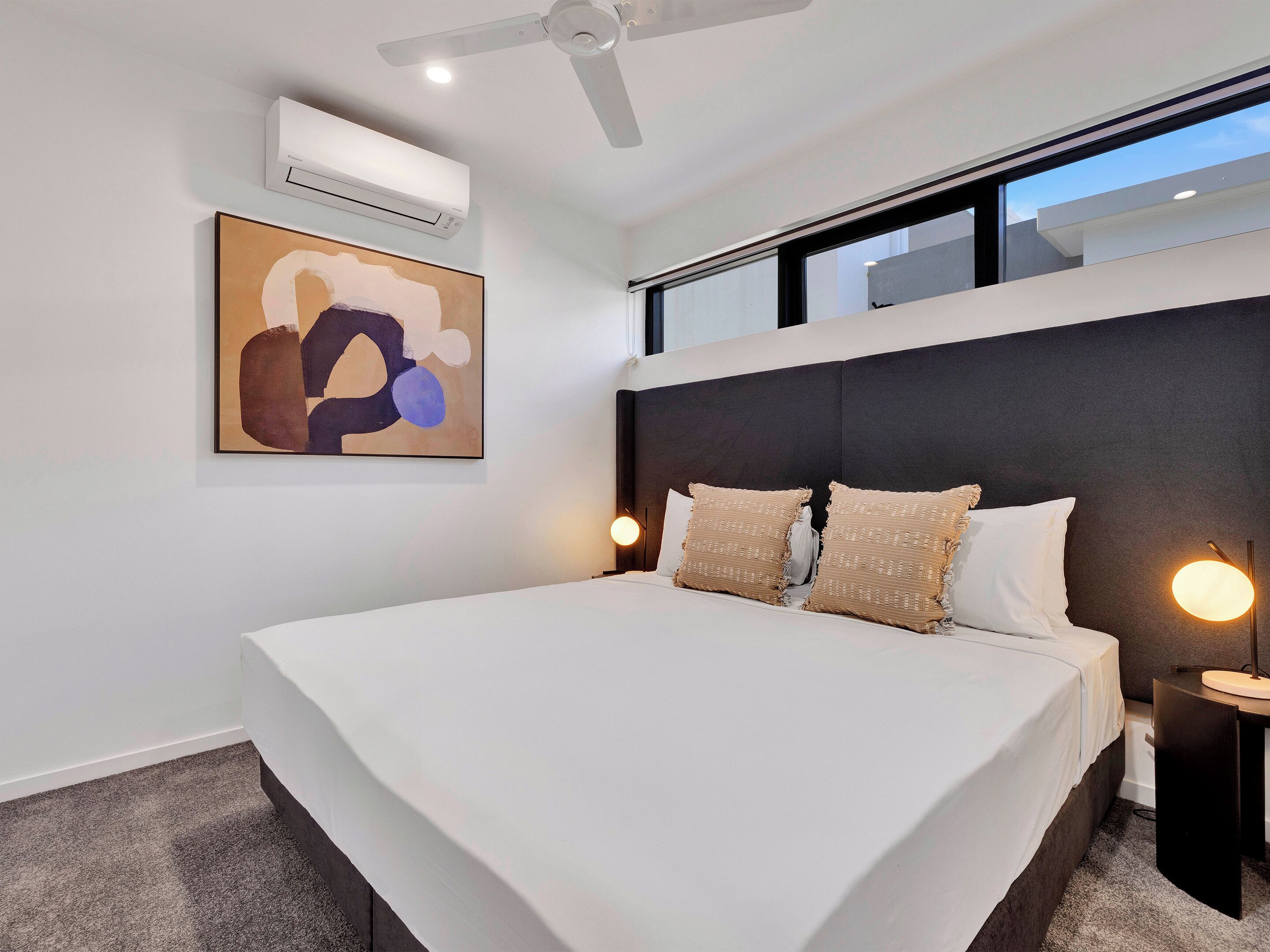 A modern bedroom with a large bed, a ceiling fan, an air conditioning unit, a wall-mounted abstract painting, and a nightstand with a lamp.