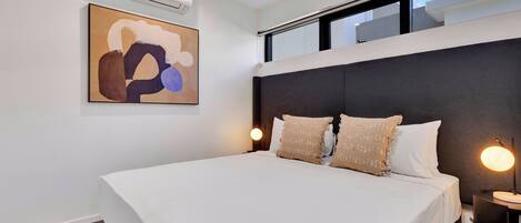 A modern bedroom with a large bed, a ceiling fan, an air conditioning unit, a wall-mounted abstract painting, and a nightstand with a lamp.