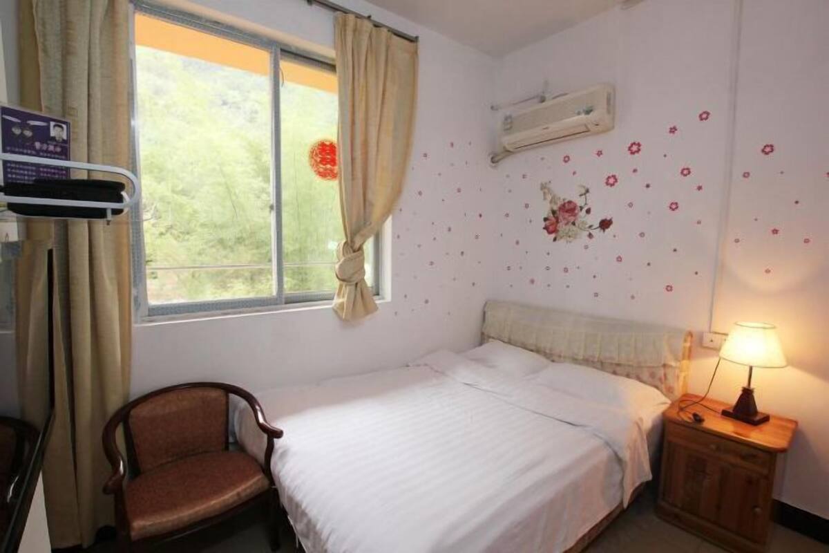 Una habitación con cama, una silla, una mesita de noche con lámpara, una ventana con cortinas y un aire acondicionado instalado en la pared.