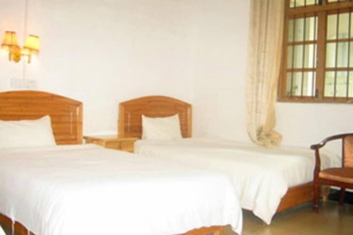 Habitación de hotel con dos camas de madera, ropa de cama blanca, una mesita de noche con lámpara y una ventana con cortinas.