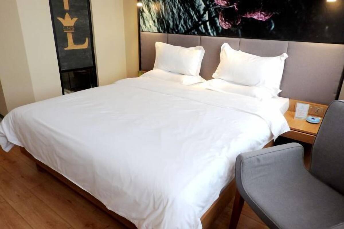 Een hotelkamer met een groot bed, een grijs gestoffeerde fauteuil, een nachtkastje met een telefoon en een muur met een logo.