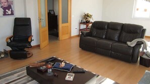 Una sala de estar con un sofá de cuero negro, un sillón negro, una mesa de centro con varios objetos y una puerta de madera que da a otra habitación.