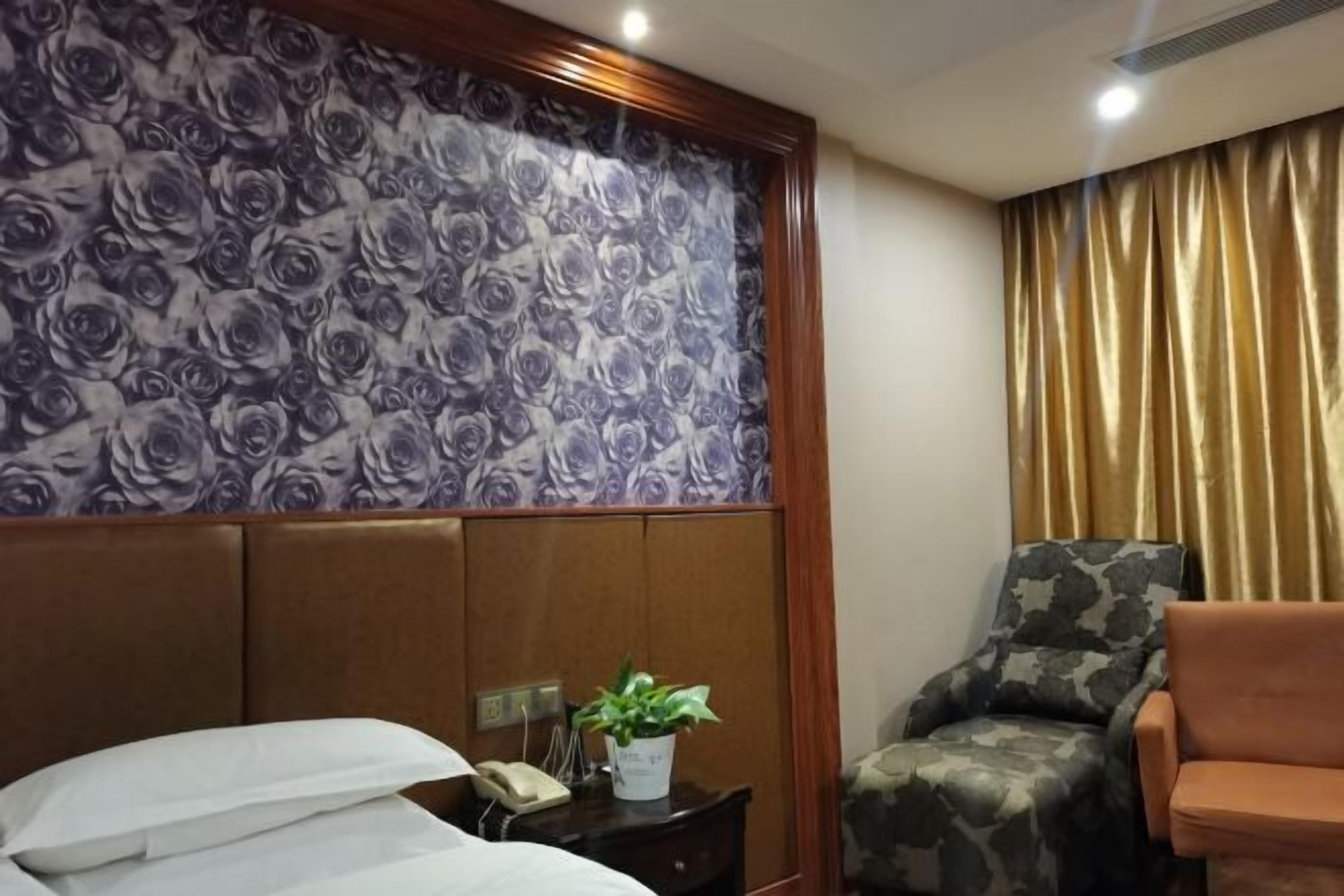 Habitació d'hotel amb paper pintat floral, un llit amb roba blanca, una tauleta de nit amb un telèfon i una planta, una cadira de camuflatge i una cortina daurada.