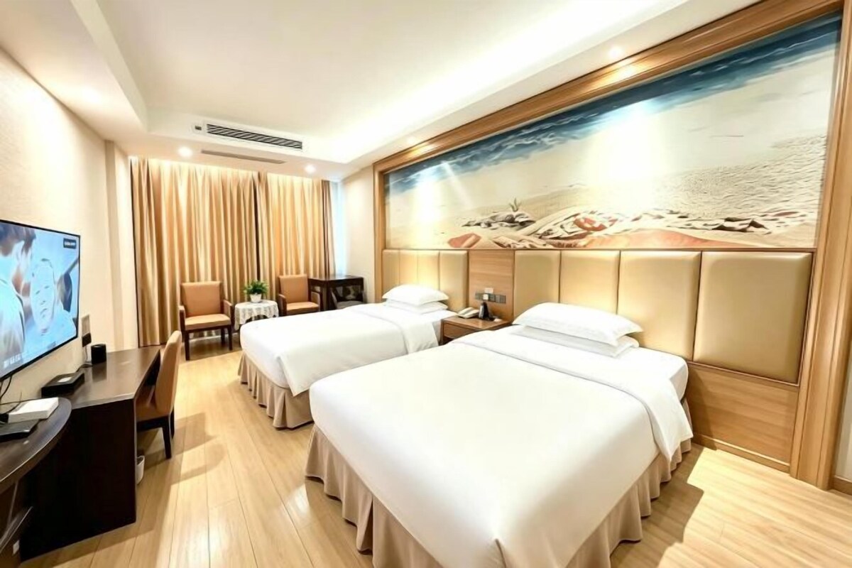 A hotel room with two beds, a desk with a TV, and a large beach mural on the wall.