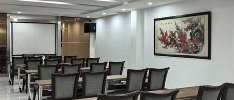 Uma sala de conferências com várias mesas e cadeiras, uma tela de projetor e uma obra de arte emoldurada na parede.