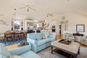A spacious living room with a light blue sofa, wooden chairs, and a patterned ottoman. A kitchen with white cabinets and a dining area with a blue rug are visible in the background. - Salty Cs: Stunning Oceanside Home with Private Pool, Hot Tub and Pool Table! (Nags Head)