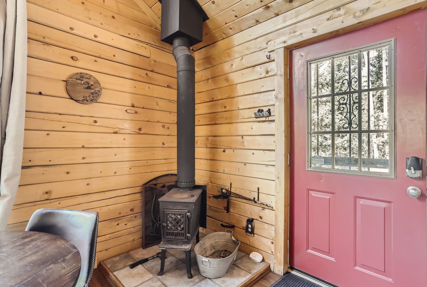 L’intérieur d’une cabane avec un poêle à bois, une porte rouge et une fenêtre avec des barreaux.