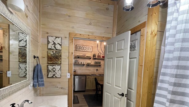 Een houten interieur met een badkamer, compleet met wastafel, spiegel en handdoekrek. Een deur leidt naar een keukenruimte met een 'GROCERY' bord.