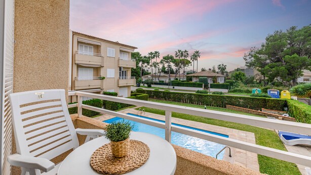 A balcony with a pool, white chairs, and a view of a residential area with palm trees and a sunset sky. - Yourhouse El Xiprer (Pollensa)