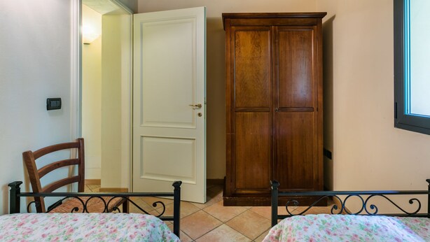 Une chambre avec une armoire en bois, un lit recouvert d’une couvre-lit à motifs floraux, une chaise et une fenêtre avec des stores.