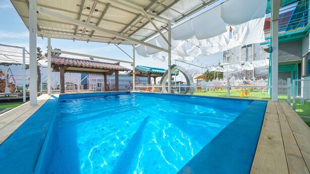 Une piscine avec une terrasse en bois, un espace couvert doté d’un toit, et un terrain de jeux clôturé avec des toboggans et des balançoires.