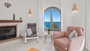 A living room with a fireplace, a grey armchair, a pink sofa, a small round table, and a view of the sea through a large window.