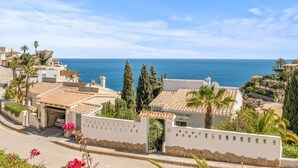 A coastal residential area with Mediterranean-style buildings, a clear blue sky, and the sea in the background.