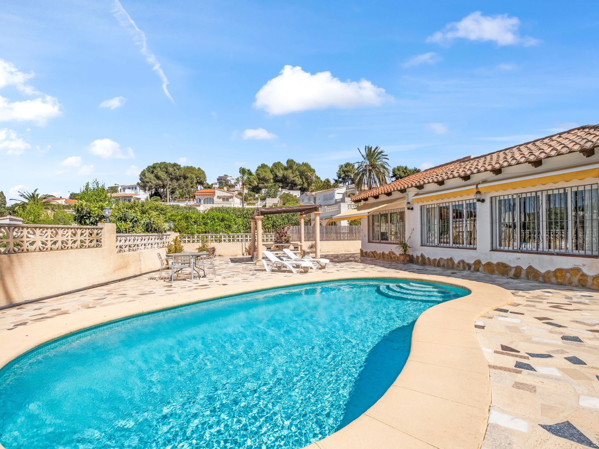 Una piscina con agua cristalina azul, rodeada por una terraza de baldosas y una pérgola.