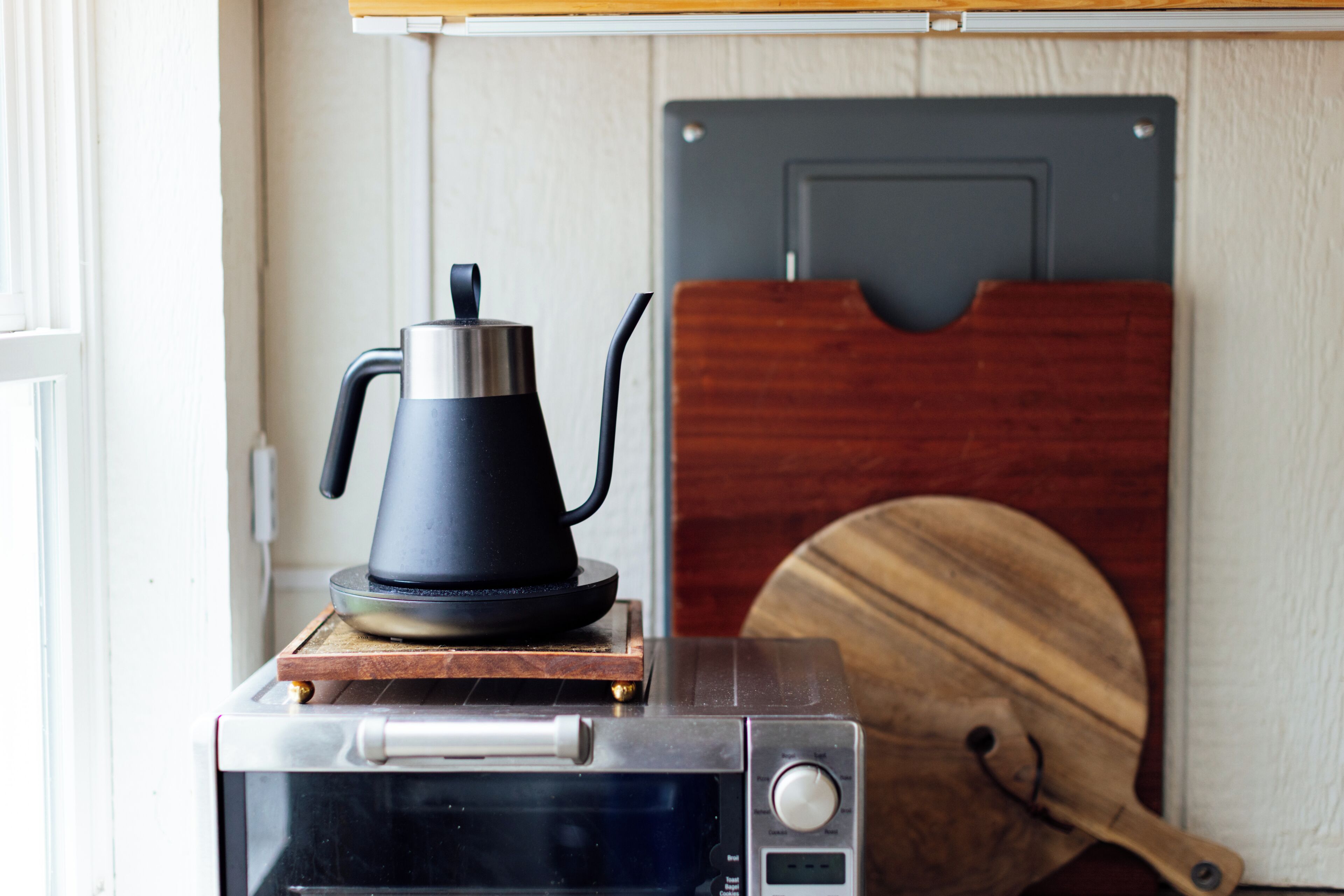 A microwave with a kettle on top, a wooden cutting board, and a door in the background.